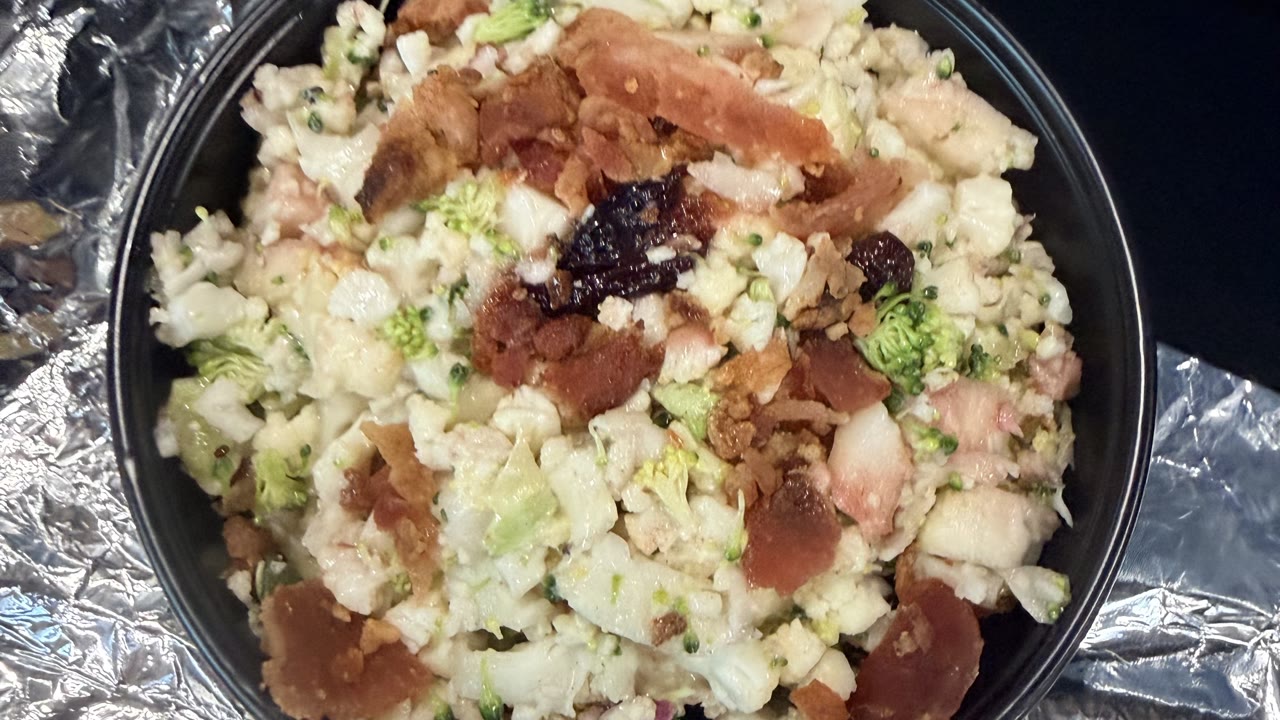 A black bowl of broccoli and cauliflower salad from above, chopped fine, with bacon, dried cranberries and a little red onion