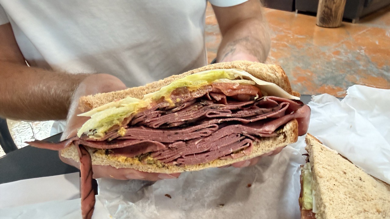 A cut half of the New Yorker held up in two hands, showing a thick fold of pastrami under lettuce, tomato and cheese