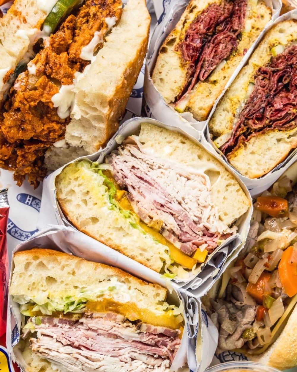 An overhead spread of Young Buck sandwiches on blue-and-white deli paper — fried chicken with pickles, two pastrami halves with mustard, and two cut clubs on thick focaccia, next to a cup of pickled vegetables